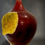 Red-glazed ceramic vase with a pale beige neck and a yellow crystalline crust attached to its side.