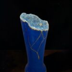 Blue ceramic vase with a jagged, crystalline blue geode top edged in gold on a dark background, decorative art piece.