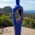 Blue ceramic vase sculpture with a purple circular opening and gold crackle pattern, set against a coastal landscape with hills and sea in the background.