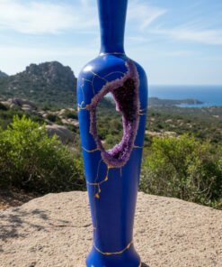 Blue ceramic vase sculpture with a purple circular opening and gold crackle pattern, set against a coastal landscape with hills and sea in the background.