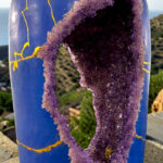 Blue glass bottle with a large purple crystalline crack/geode spilling down its side, outdoors by a hillside sea view.