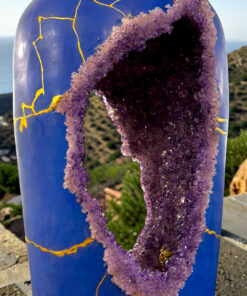Blue glass bottle with a large purple crystalline crack/geode spilling down its side, outdoors by a hillside sea view.