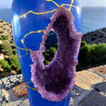 Blue glass bottle with a jagged purple amethyst geode growing through a hole, set against a coastal landscape with the sea in the background.