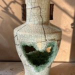 Crackled ceramic vase with two dark brown handles and a large broken opening near the lower body, revealing green crystalline interior.