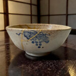 Close-up of a ceramic bowl with a blue decorative pattern and a few strings resting on a dark wooden table.