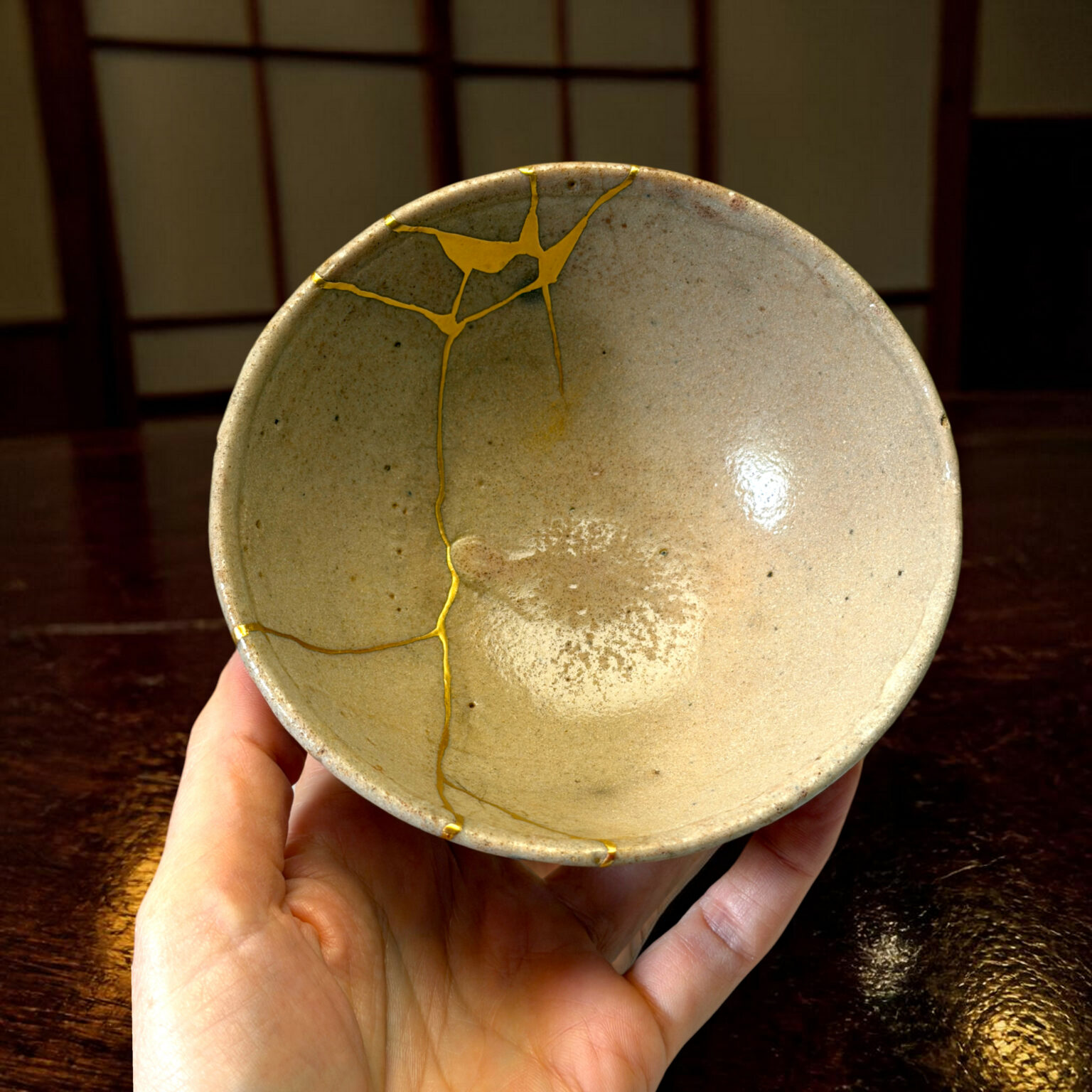 Close-up of a gray ceramic bowl repaired with gold lines, held in a hand over a dark wooden surface.