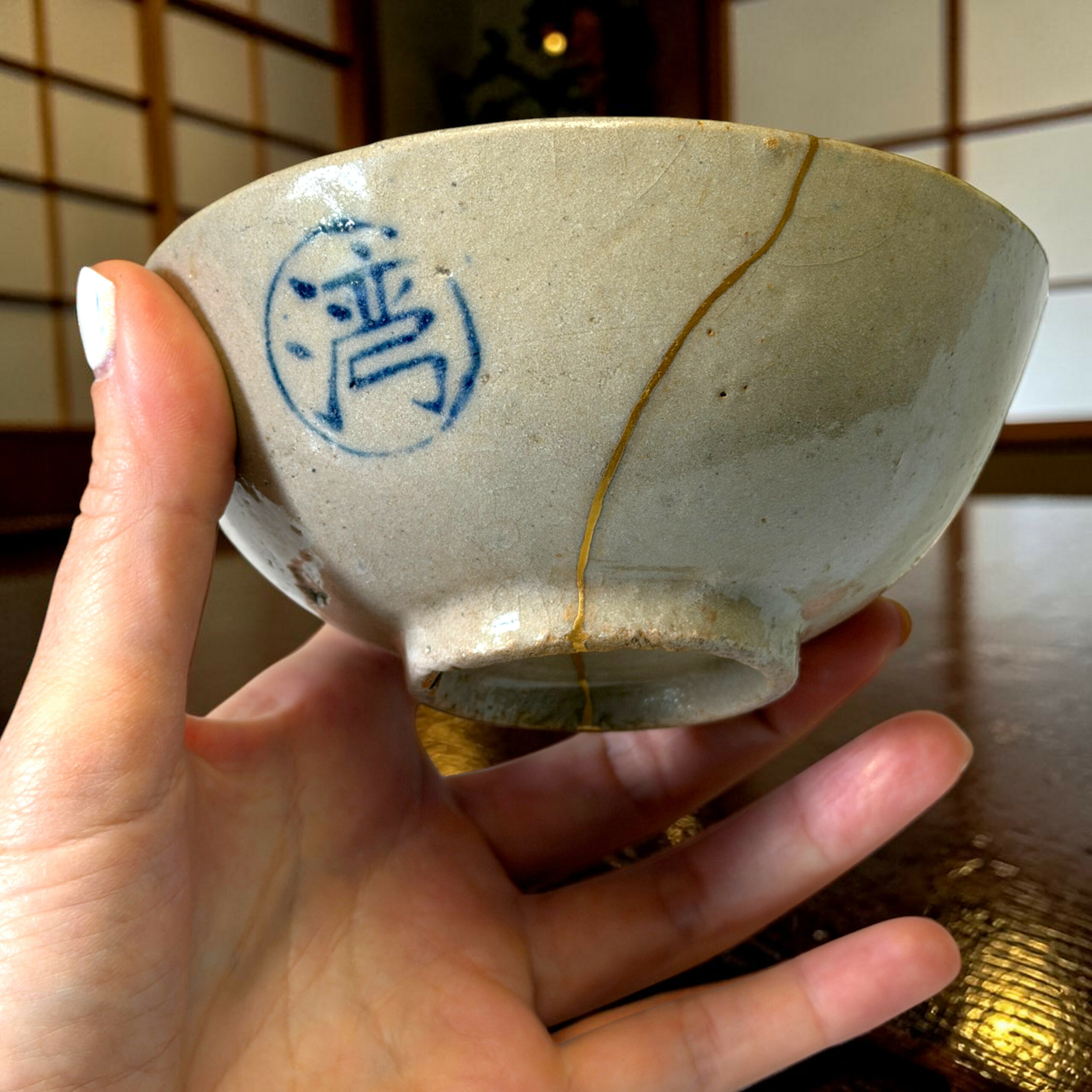 Close-up of a gray ceramic bowl with a blue circular maker's mark, held by two fingers; visible crack along the glaze and a yellow seam running down the side, background of a Japanese-style room.