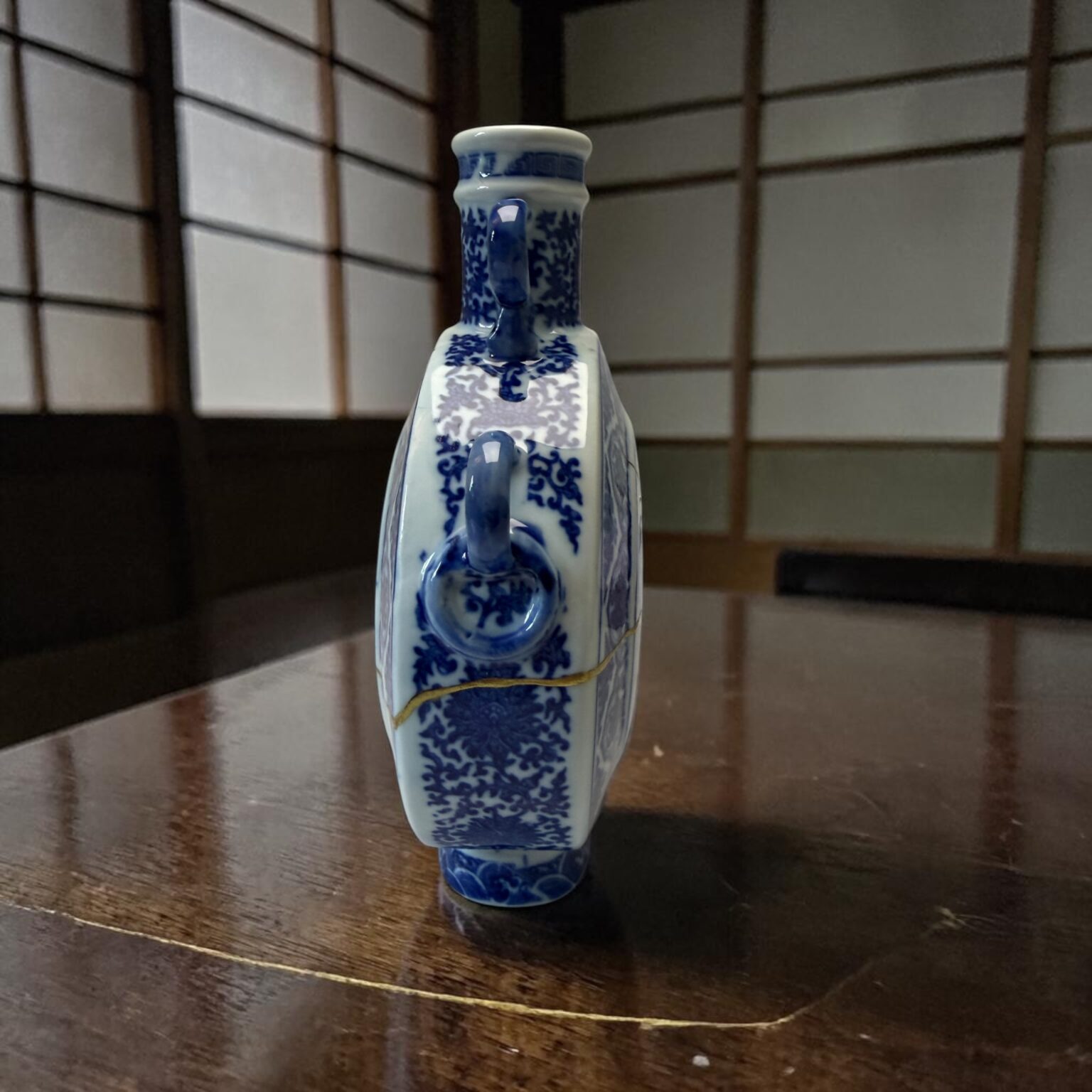 Blue-and-white porcelain vase with intricate floral patterns sits on a polished wooden table, background showing grid-panel walls.