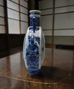 Blue-and-white porcelain vase with intricate floral patterns sits on a polished wooden table, background showing grid-panel walls.