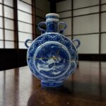 Blue and white porcelain vase with ornate patterns sits on a dark wooden table against a grid-pattern wall.