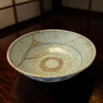 Close-up of a pale blue ceramic plate with gold kintsugi repair lines, set on a dark wooden table.