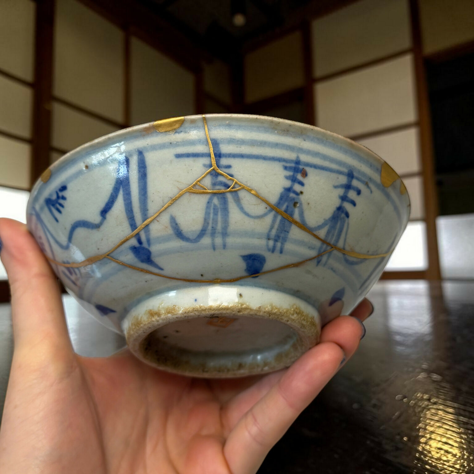 Close-up of a blue-and-white ceramic bowl repaired with gold kintsugi seams, held by a hand in the foreground.