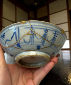 Close-up of a blue-and-white ceramic bowl repaired with gold kintsugi seams, held by a hand in the foreground.