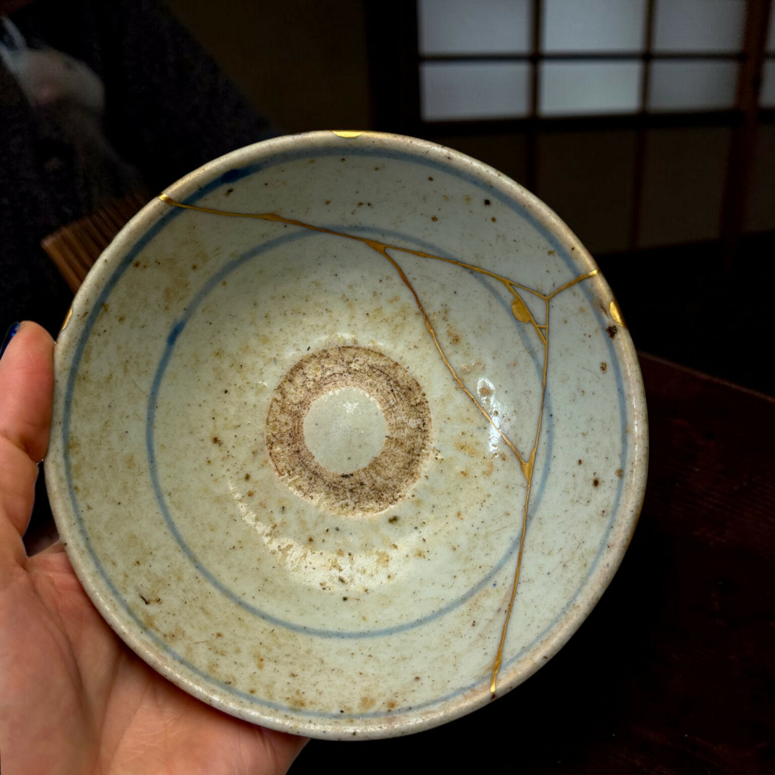 Close-up of a light-blue ceramic plate with gold crackle lines, held by a hand, against a dark background and a blurred window in the back.