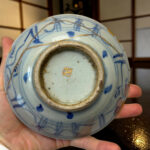 Close-up of a pale blue porcelain vase being held, showing blue brushwork, a dirty inner rim, and gold crack lines along the edge.