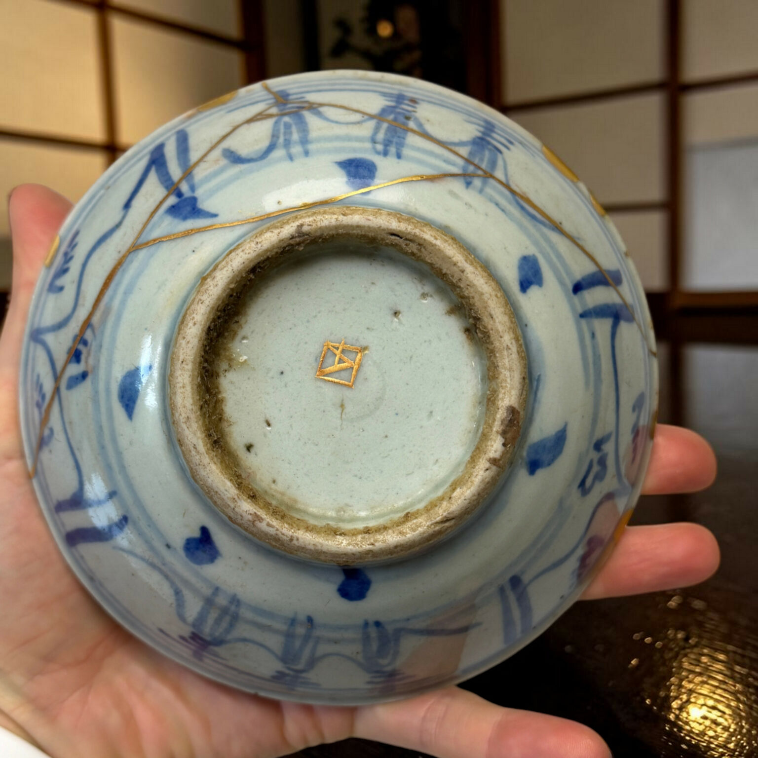 Close-up of a pale blue porcelain vase being held, showing blue brushwork, a dirty inner rim, and gold crack lines along the edge.