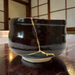 Close-up of a glossy black ceramic mug with a gold crack running vertically down the side, resting on a wooden table.