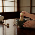 Hands whisking powdered tea in a ceramic bowl with a bamboo chasen during a matcha ritual.