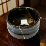 Close-up of a dark ceramic cup with gold tape along its rim, sitting on a wooden table.