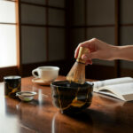 Hand whisking matcha with a bamboo chasen over a black ceramic bowl on a wooden table; open book, cup, and sifter nearby in a calm setting