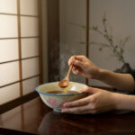 Hands hold a ceramic bowl of steaming soup as a wooden spoon stirs inside.