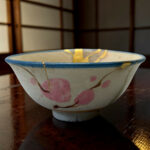 Close-up of a ceramic bowl with a blue rim and pink floral designs, placed on a dark wooden table with a blurred wooden screen in the background.