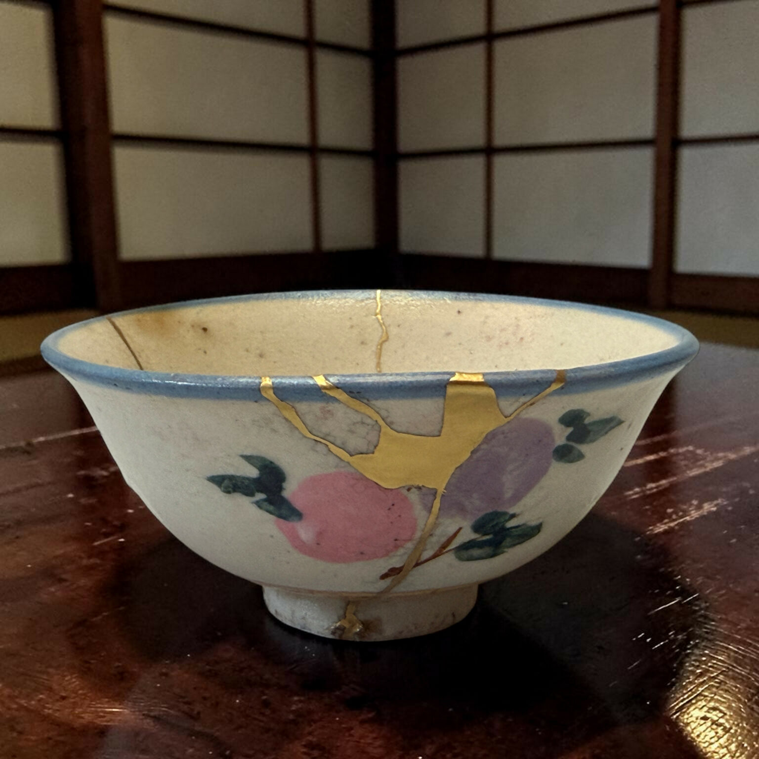 Close-up of a ceramic bowl with gold kintsugi repair lines and a pink/green floral pattern on a dark wooden table.