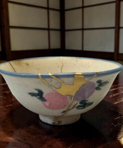Close-up of a ceramic bowl with gold kintsugi repair lines and a pink/green floral pattern on a dark wooden table.