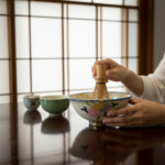 Hands whisking matcha in a decorative ceramic bowl with a bamboo whisk on a dark wooden table.