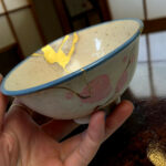 Close-up of a small ceramic bowl with a blue rim, yellow decoration, held in a person's hands.