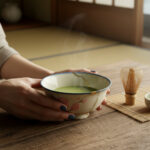 Hands cradle a ceramic bowl of steaming green matcha on a wooden table with a bamboo whisk nearby.