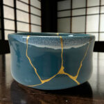 Blue ceramic mug with gold decorative lines sits on a dark wooden table; background features a blurred grid pattern.