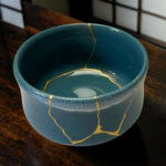 Blue ceramic mug with gold repair lines (kintsugi) running along the rim and interior, placed on a wooden table.