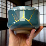 Close-up of a teal ceramic mug with gold crackle lines being held by a hand, background is blurred.