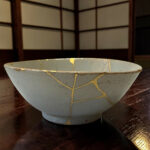 Gray ceramic bowl with yellow tape repair lines on a dark wooden table. Background shows a wooden cabinet with panels.