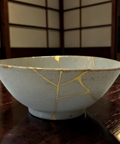 Gray ceramic bowl with yellow tape repair lines on a dark wooden table. Background shows a wooden cabinet with panels.