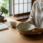Person gently holding a pale ceramic bowl at a wooden table, with a steaming cup of tea and an open book nearby.