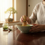 Person whisking matcha in a ceramic bowl on a wooden table, with a jar of matcha powder nearby.