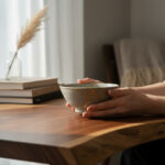 Hands cradle a speckled ceramic bowl on a wooden table, with a stack of books and a glass vase in the softly lit background.