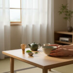 Hands cradle a pale green bowl on a light wooden table, with a teapot, small bowl, whisk, and leaves nearby in a sunlit room.