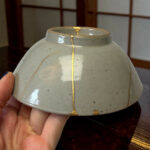 Close-up of a gray ceramic vase with gold kintsugi repairs, being held by a person’s fingers.