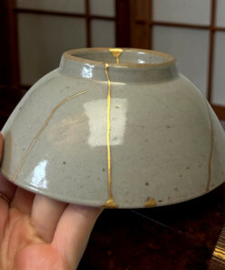 Close-up of a gray ceramic vase with gold kintsugi repairs, being held by a person’s fingers.