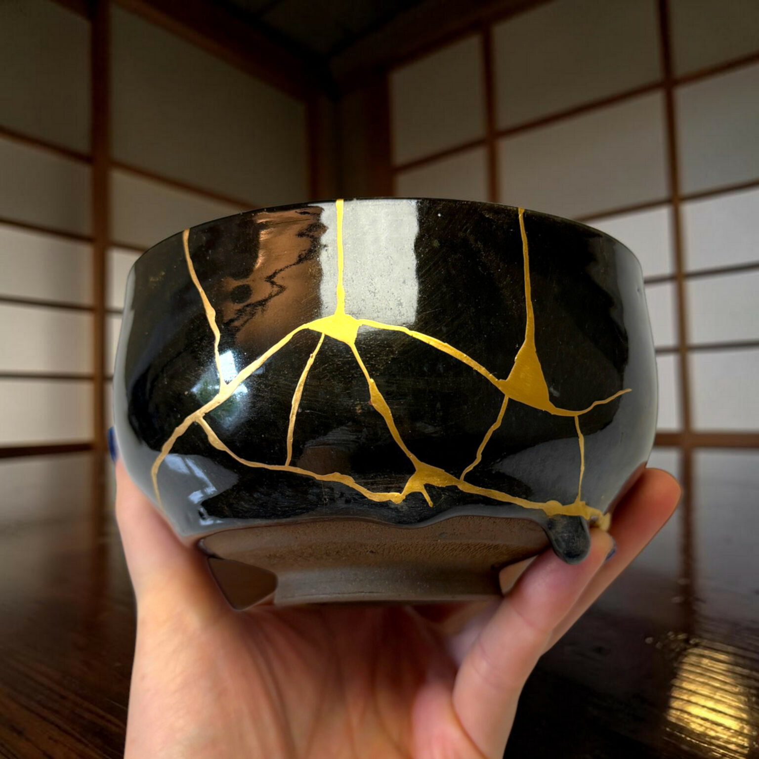 Close-up of a glossy black ceramic vessel with gold crackle pattern being held by hands, with wooden slats in the background.