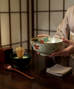 Person in traditional attire holds a decorative Japanese tea bowl over a dark wooden table with a green matcha bowl and whisk nearby, in a setting with shoji screens behind.