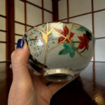 Close-up of a decorated ceramic vase being held by a hand with blue nail polish, showing gold crackle seams and leaf patterns.