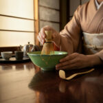 Person in a traditional kimono whisks matcha tea with a bamboo chasen over a cracked green tea bowl at a Japanese-style setup.