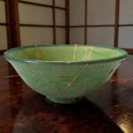 Close-up of a green ceramic bowl with gold decorative lines resting on a dark wooden surface, lattice backdrop in the background.