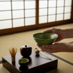 Hands holding a green ceramic tea bowl in a Japanese tea ceremony setup on a wooden tray with utensils.