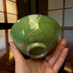 Close-up of a green sphere with gold vein-like cracks being held in both hands over a wooden surface.