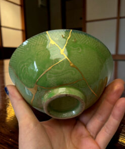 Close-up of a green sphere with gold vein-like cracks being held in both hands over a wooden surface.